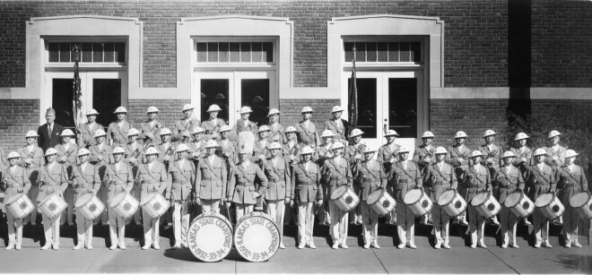 American Legion members in band uniforms stand with their instruments in front of a brick building.