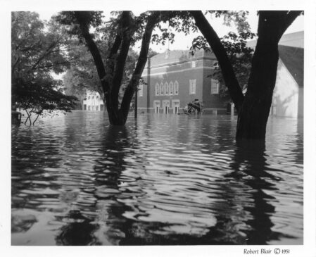 A brick building with stone accents is framed by two trees. Water stretches across the image; the building is partially submerged.