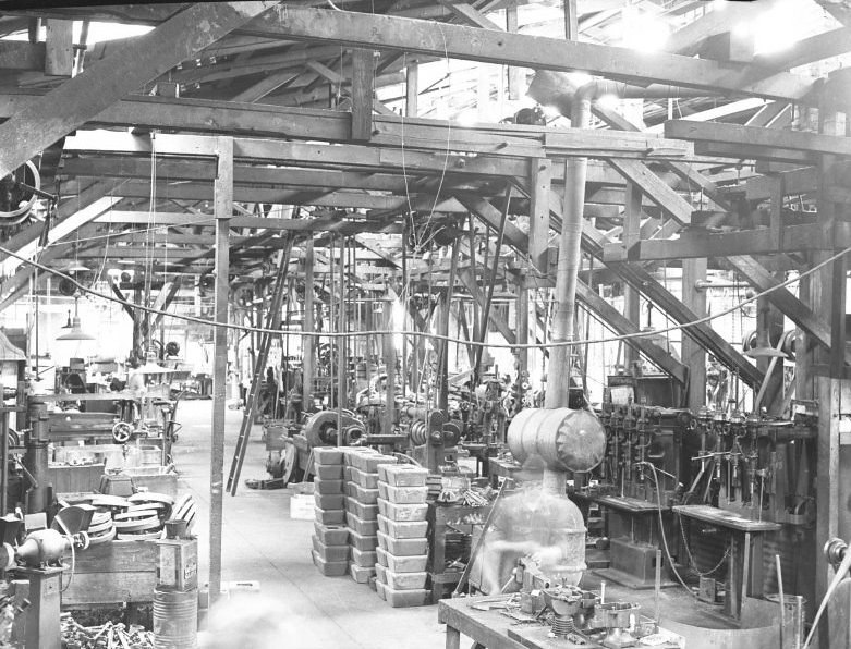 Interior of an industrial manufacturing plant. Image includes an overhead belt and numerous metal components.